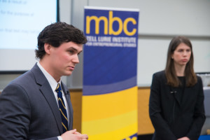 Serious, well-dressed Ross Business School student stands to the left of the Michigan Business Challenge logo with hands folded in front of him
