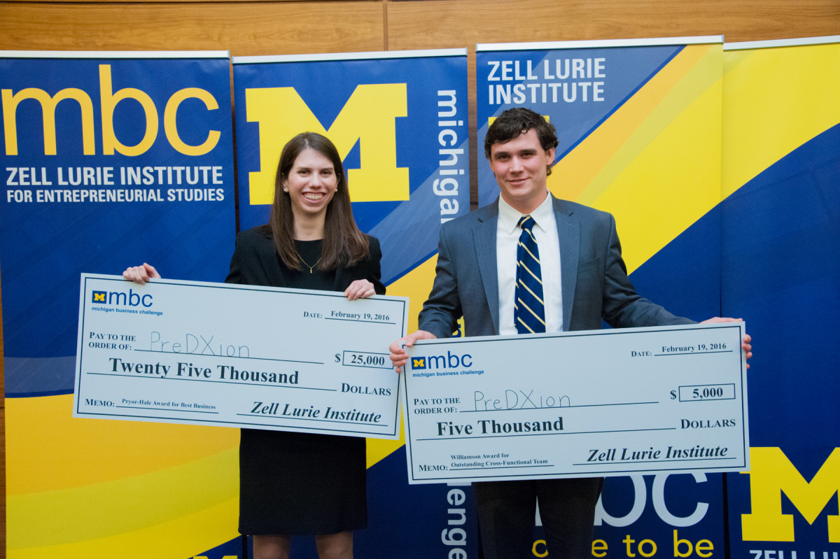 Two smiling Michigan Business Challenge contestants holding two large checks. The check in the young woman on the left's hands reads, "Pay to the order of PreDXion, $25,000 Twenty Five Thousand Dollars, Zell Lurie Institute." The check in the young man on the right's hands reads, "Pay to the order of: PreDXion, $5,000 Five Thousand, Zell Lurie Institute."